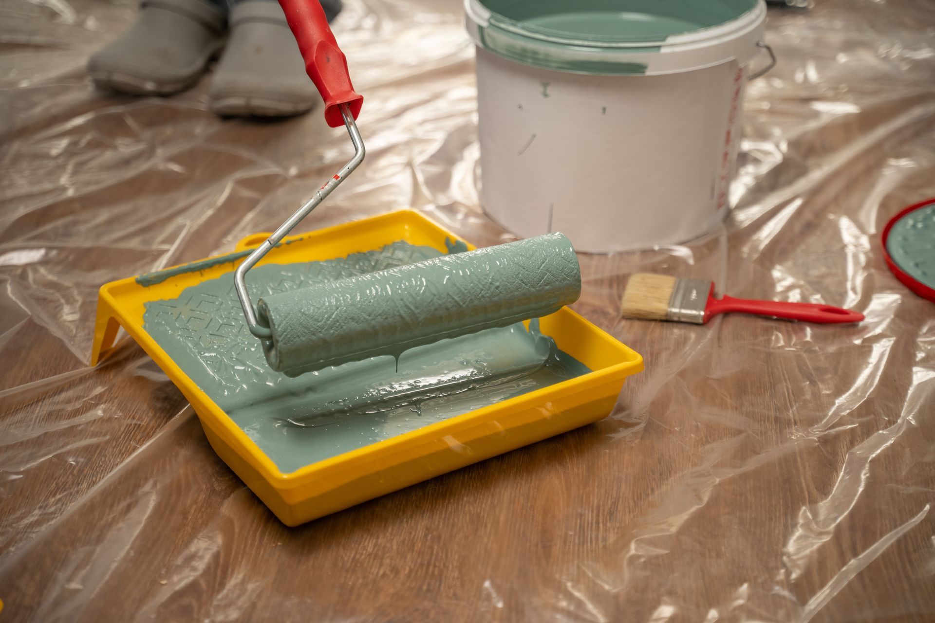 Paint roller in a yellow tray with gray-green paint beside a white paint bucket on a drop cloth