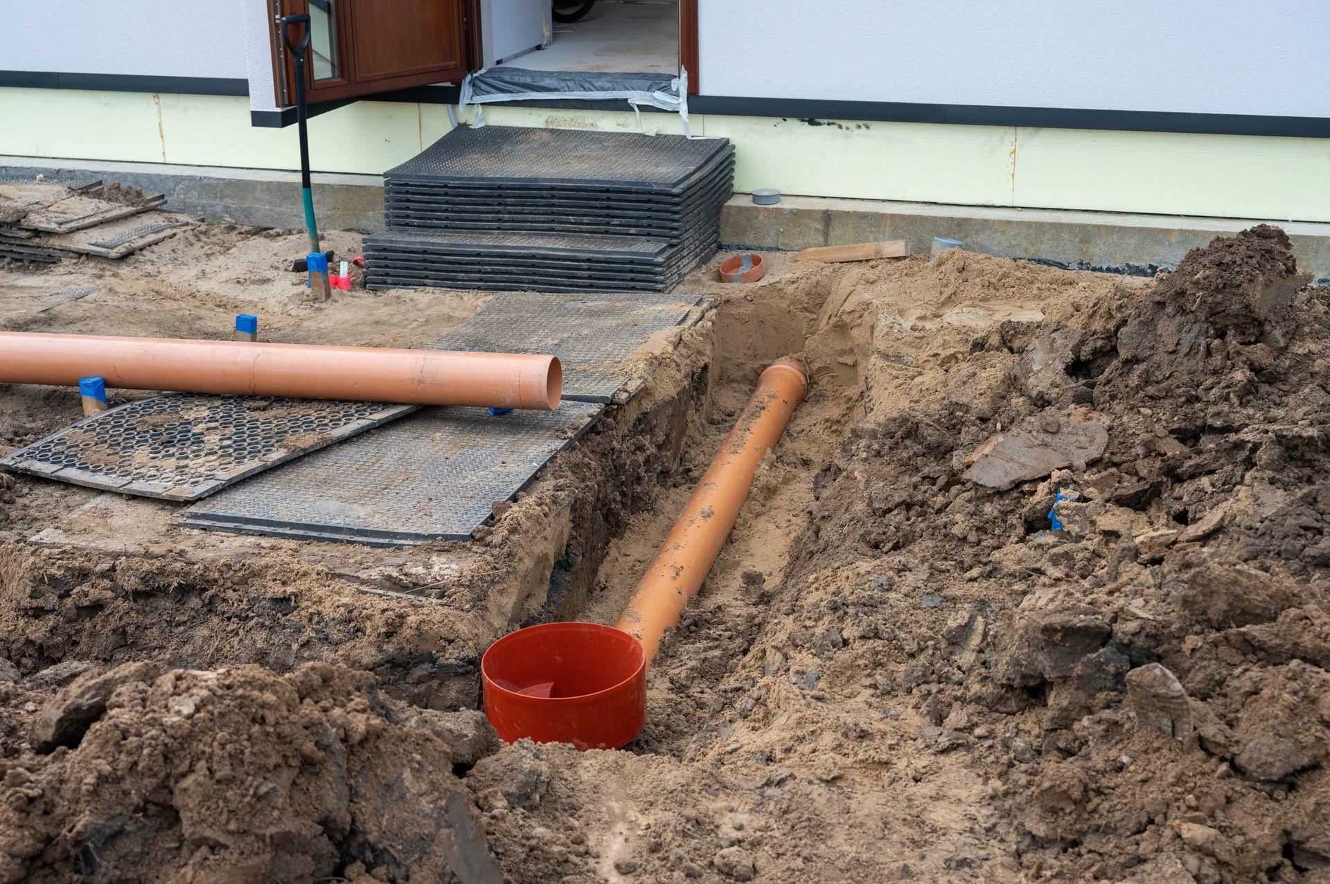Construction trench with orange PVC pipes and stacked materials beside a house foundation