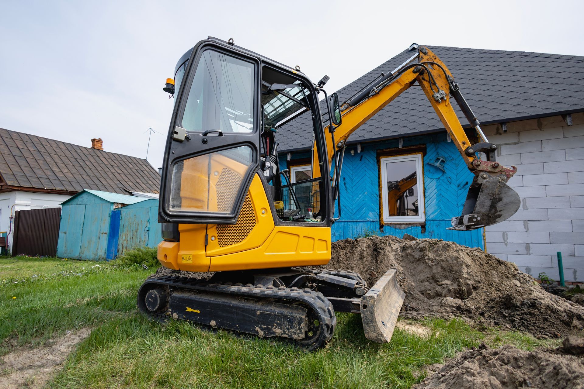 Yellow excavator demolishing a blue house wall in a grassy yard