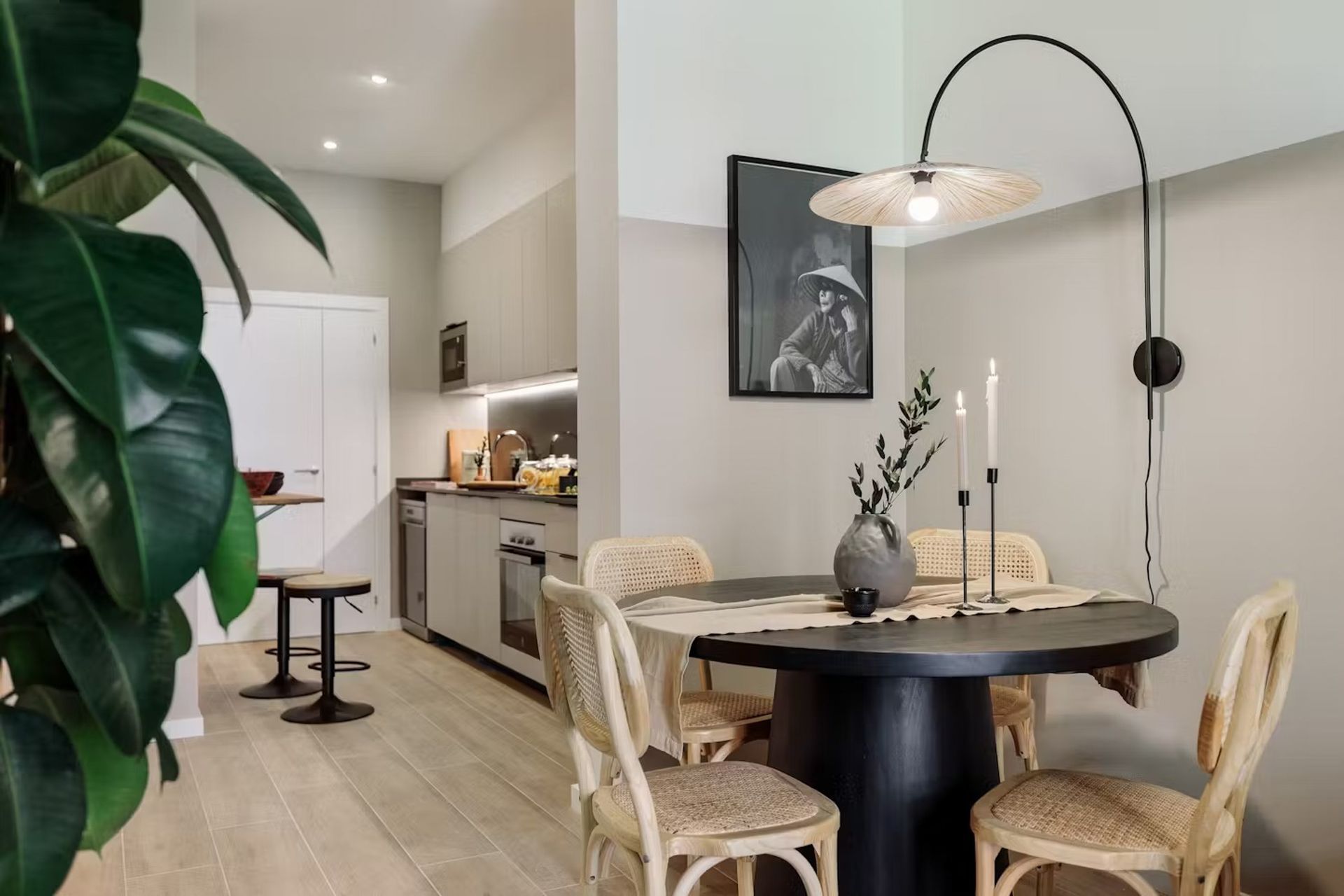 Modern dining nook with round black table, woven chairs, arched lamp, and a view into a compact kitchen.