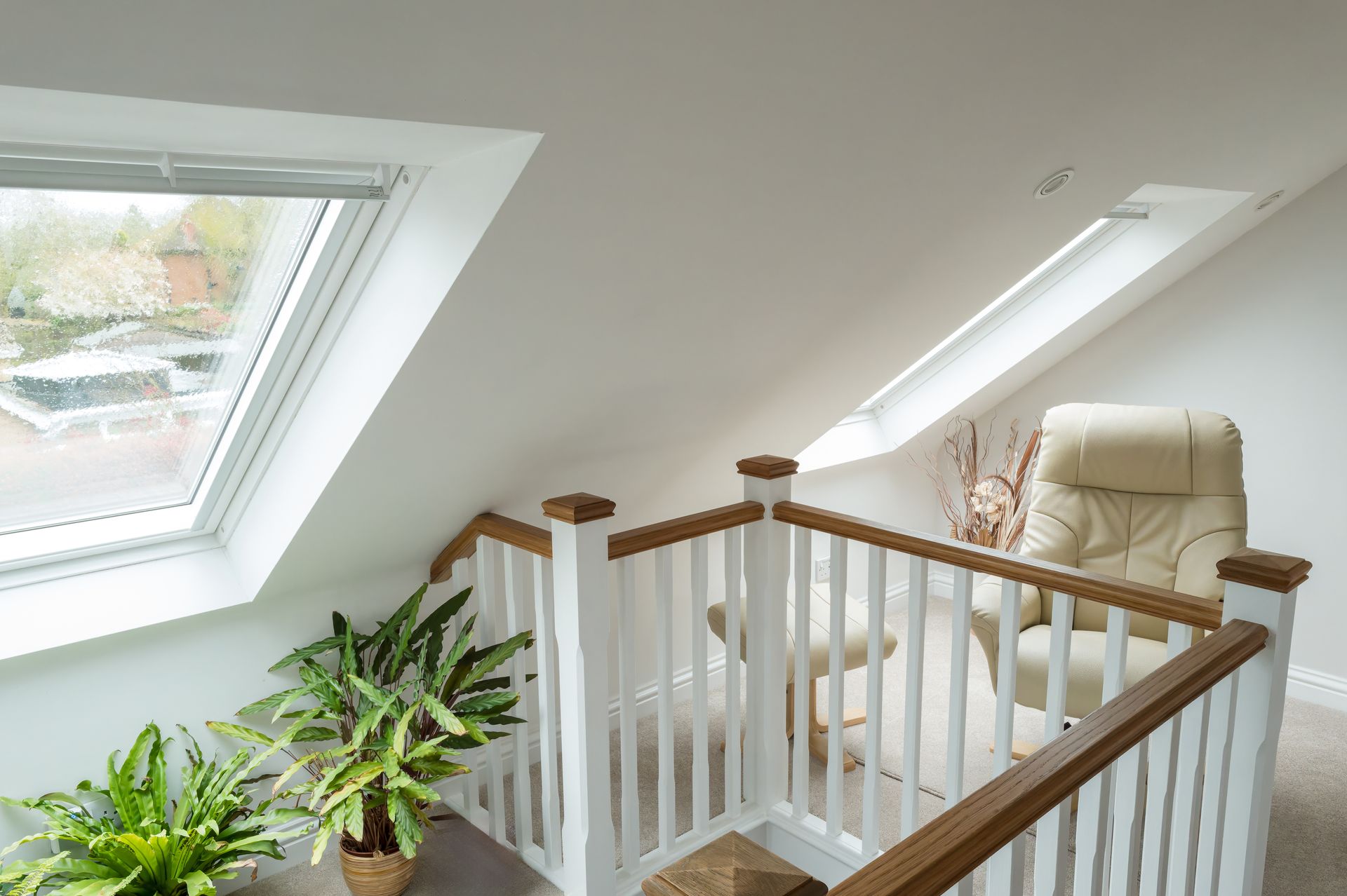 Bright attic stair landing with skylights, white railing, potted plants, and a cream chair.