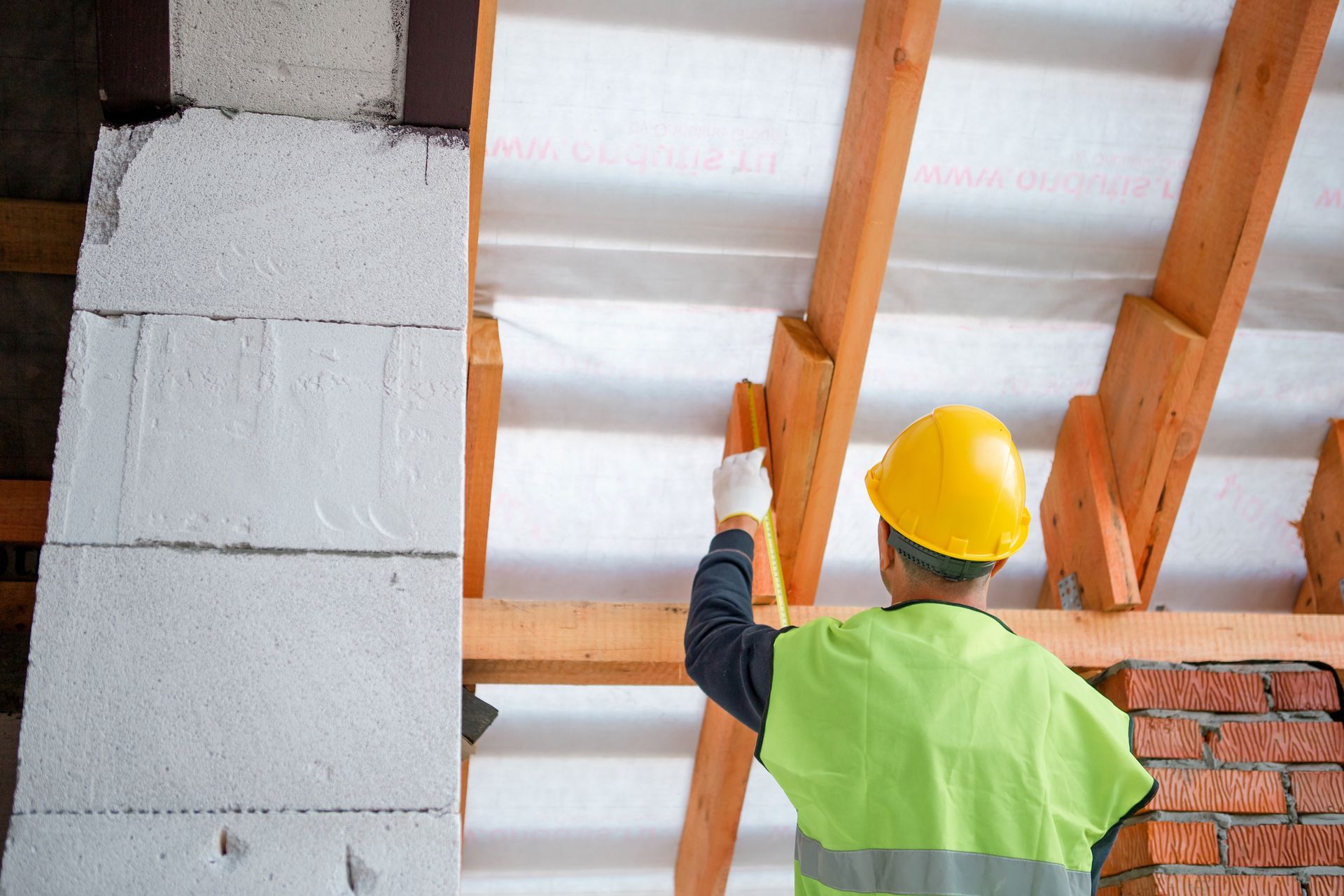 Worker in yellow hard hat inspecting roof insulation and wooden beams inside a building under construction