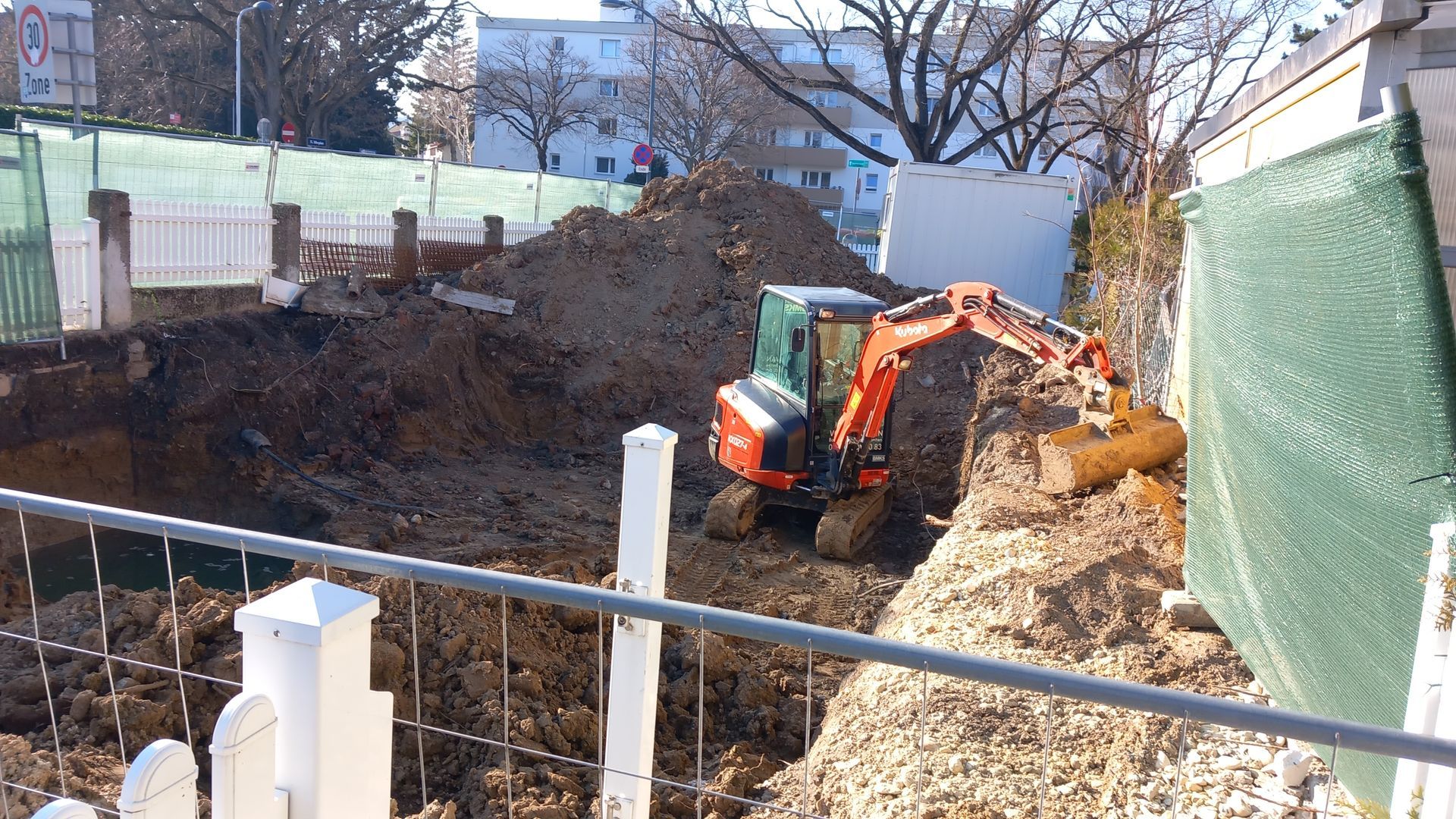 Excavator digging a large trench beside a green fence in a backyard construction site