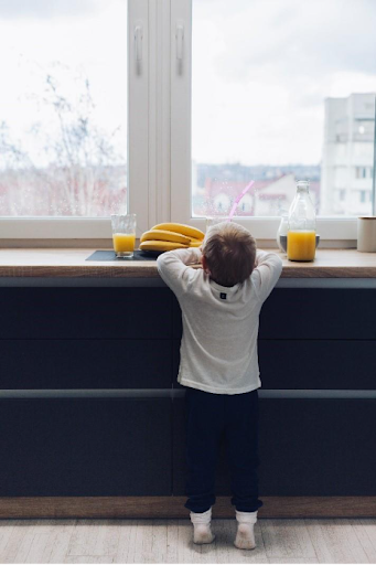 A toddler stands on toes, looking out a window from a kitchen counter; bananas and juice on the sill.