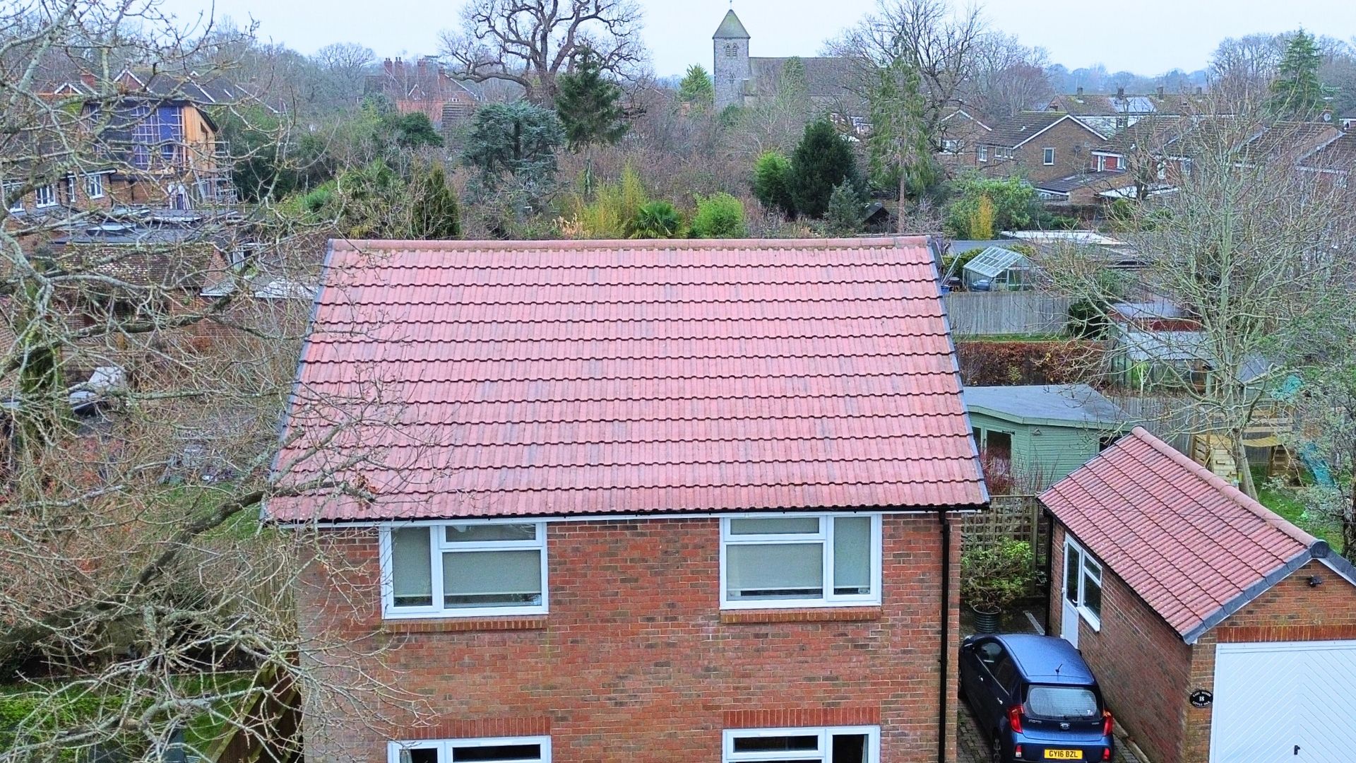 Brick house with a red-tiled roof, two windows on each floor, a car parked in the driveway, and a church in the background.