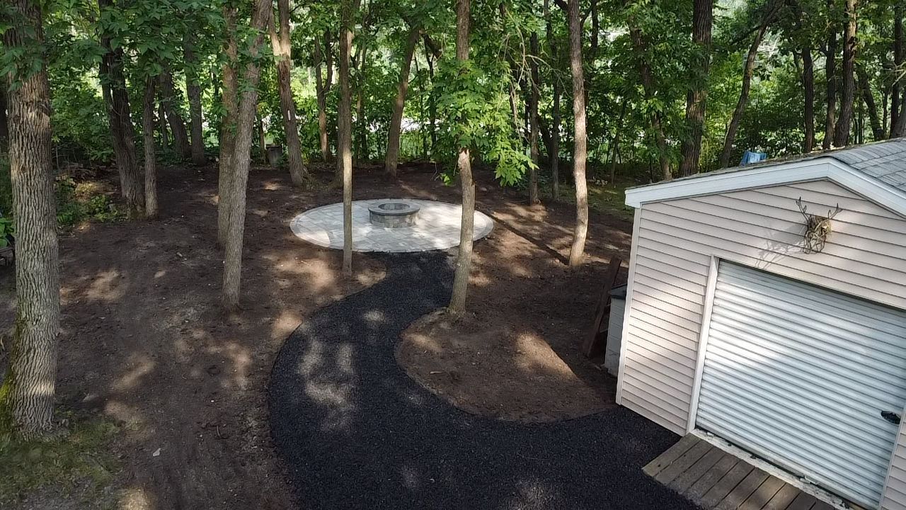 A gravel path leads to a circular fire pit area in a wooded backyard, situated next to a light-colored storage shed.