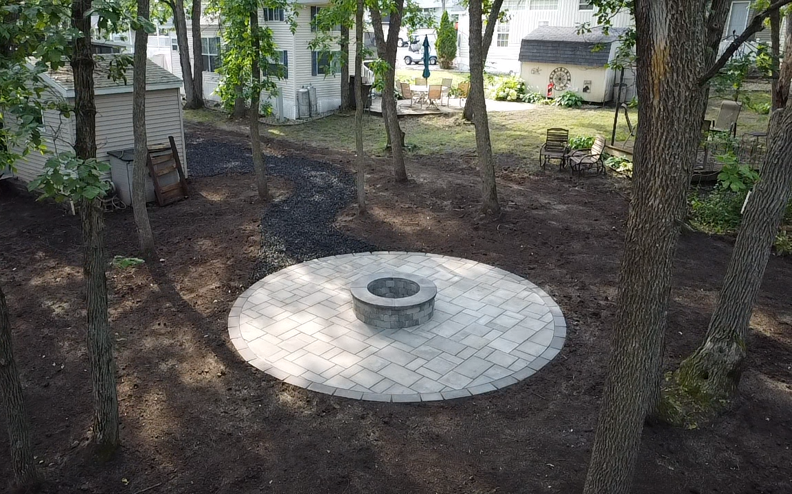 A circular stone paver patio with a stone fire pit in the center, situated in a wooded backyard near a small shed.