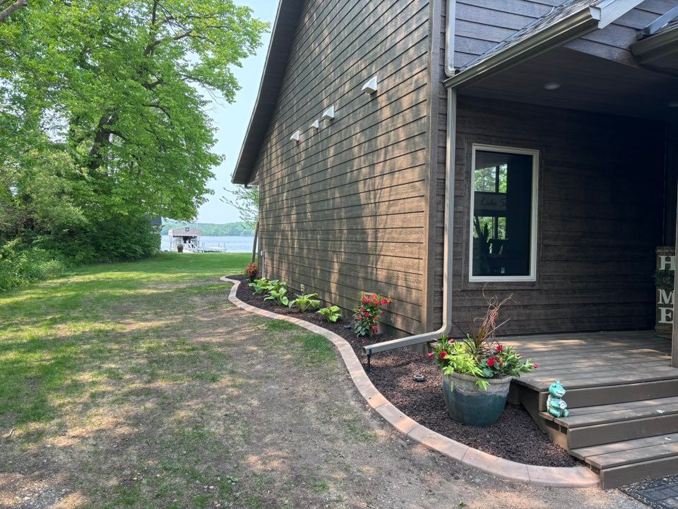 A dark wood house exterior with a curved garden bed featuring small green plants, mulch, and a potted plant by the steps.