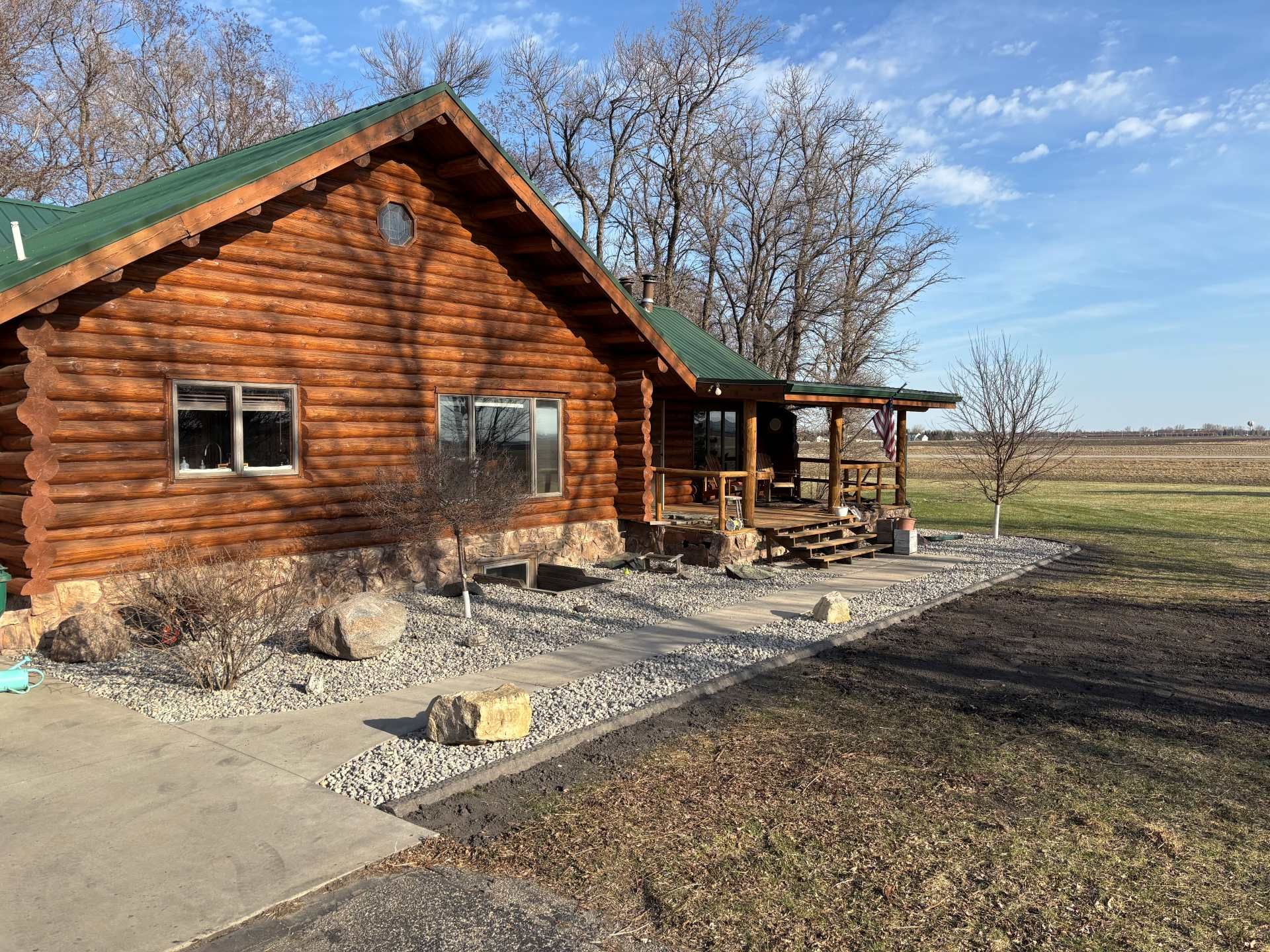 A rustic log cabin with a green metal roof, set against a backdrop of bare trees and a flat, open field on a sunny day.