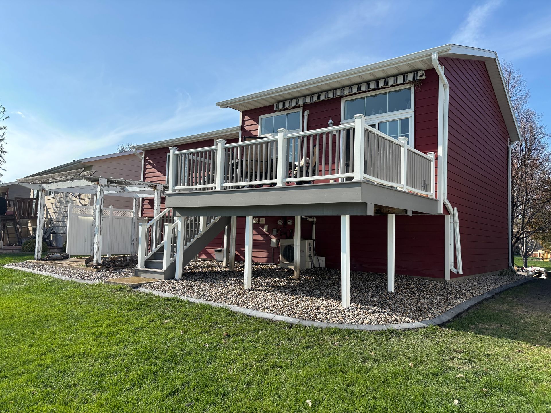 A red house with a raised wooden deck, white railings, and a side pergola over a gravel-landscaped yard under a blue sky.