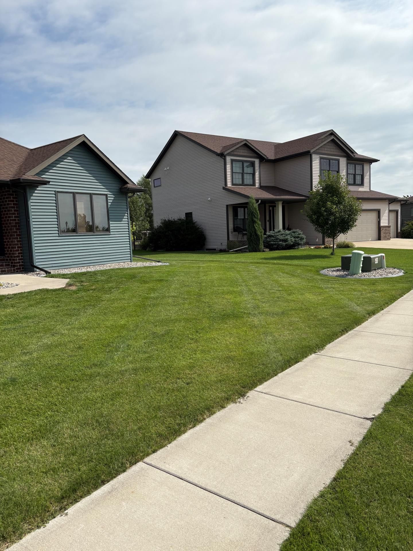 A view of a grey house and a smaller, blue-sided structure from a sidewalk next to a green lawn under a cloudy sky.