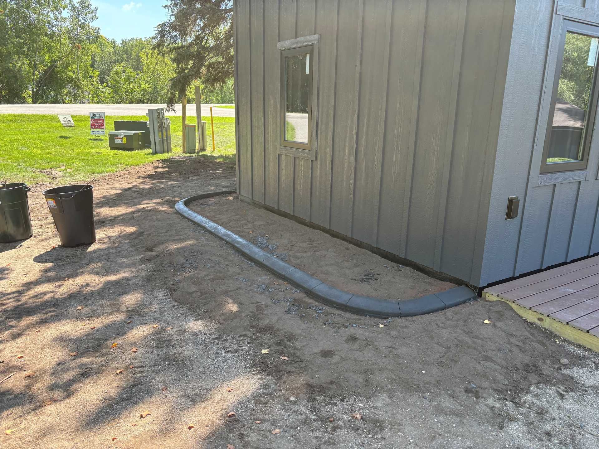 A modern gray building with vertical siding features a curved concrete landscape border edging around a dirt garden bed.
