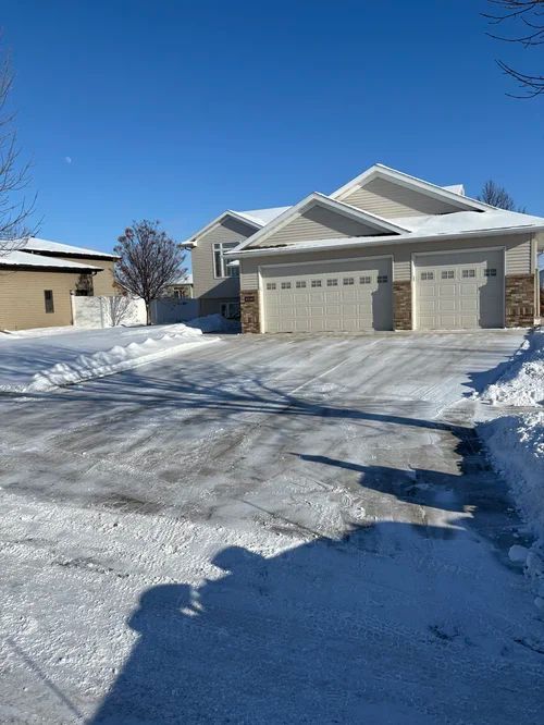 Snowy suburban house with dark siding, two-car garage, and blue recycling bins at dusk