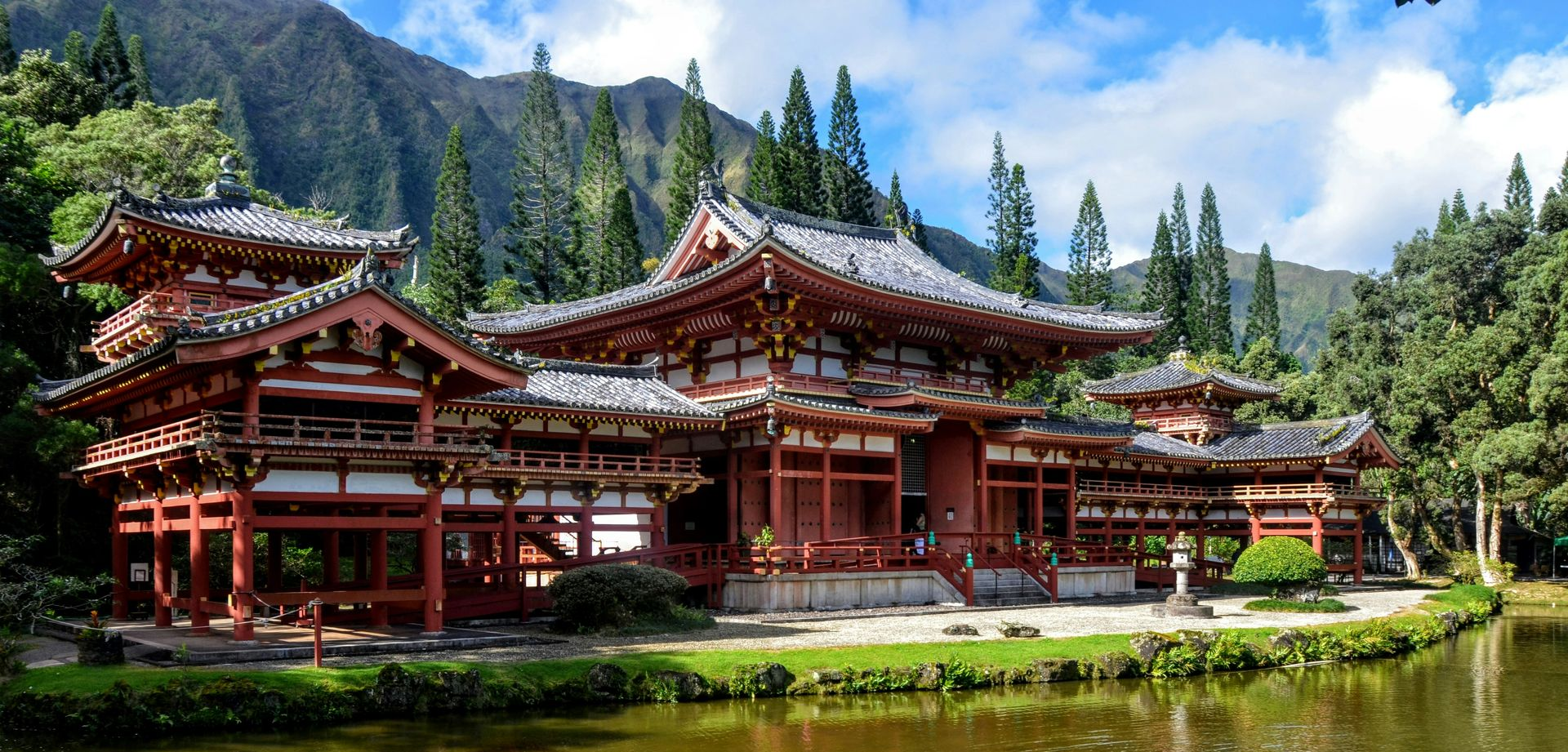 Traditional red temple complex beside a calm pond, with mountains and pine trees in the background