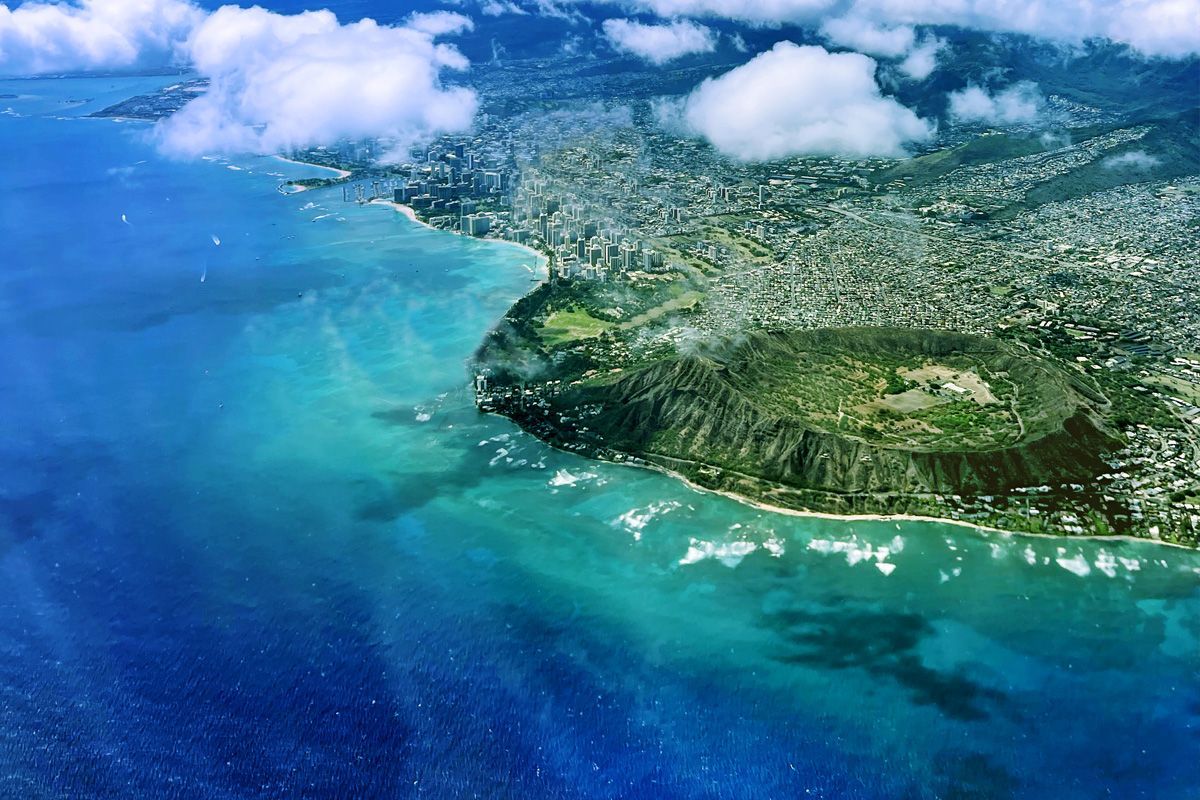 Aerial view of Diamond Head crater on Oahu, Hawaii, with the city of Honolulu behind it. Blue ocean surrounds the land.
