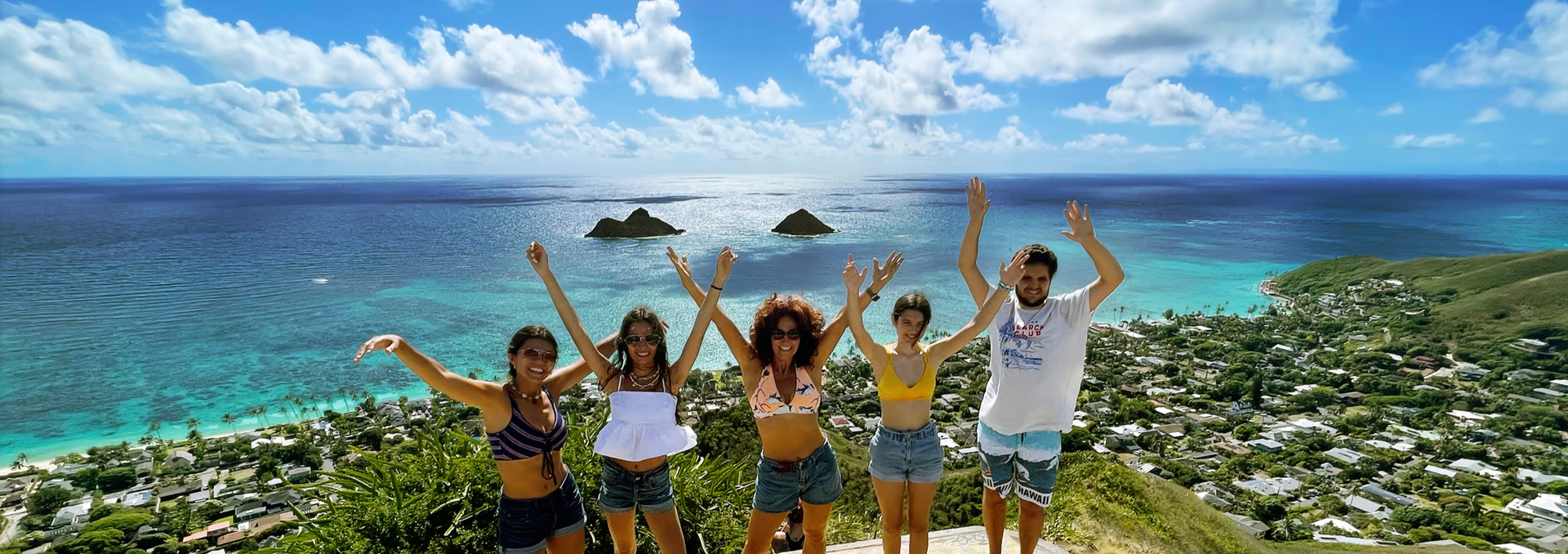 Five people stand on a hill overlooking a tropical beach and town, smiling with arms raised against a blue, cloudy sky.