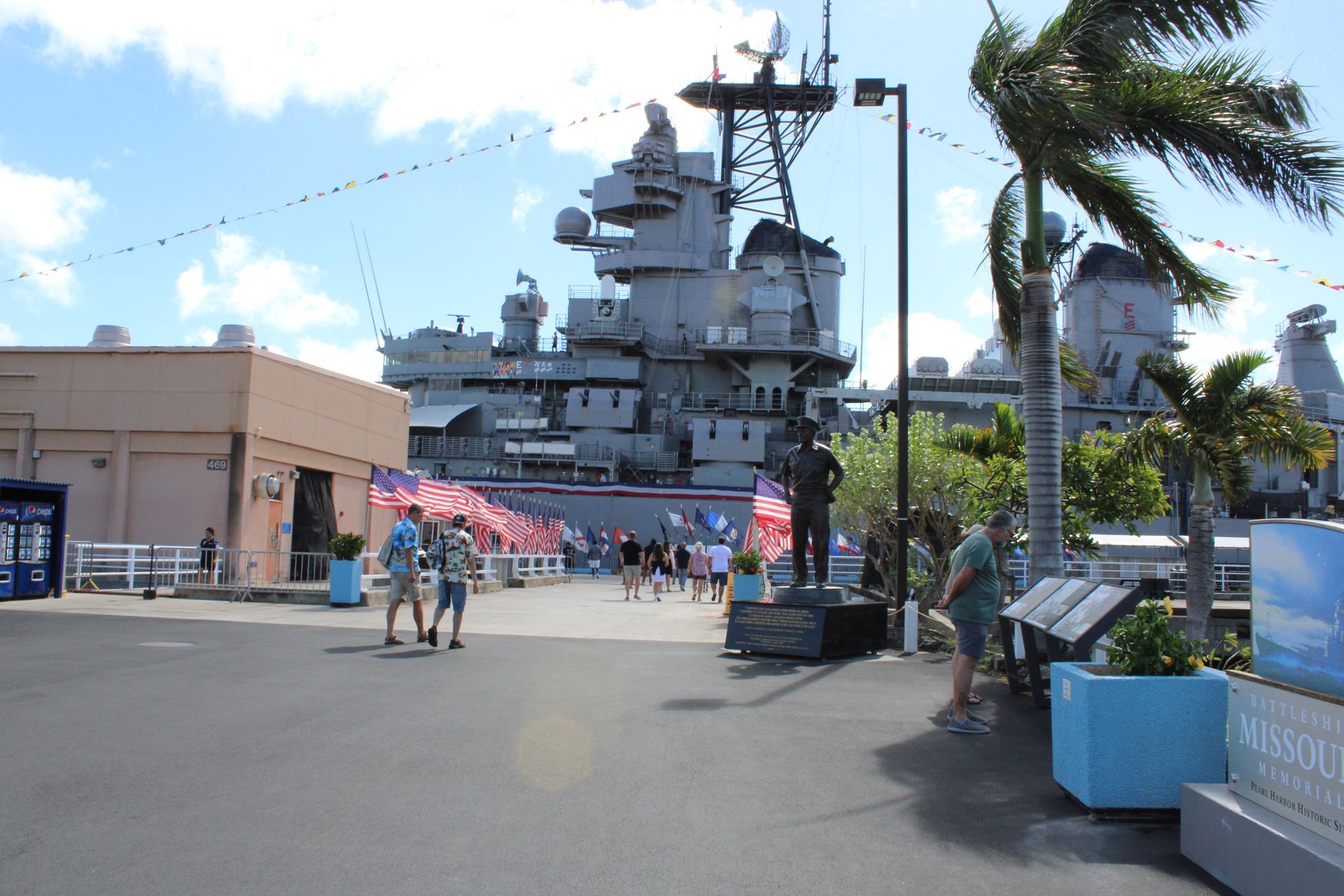 People walking towards a battleship, possibly a memorial. Sunny day. Flags and palm trees.