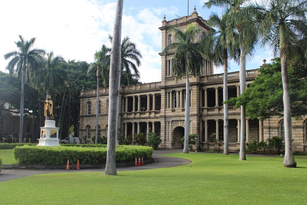 Historic colonial-style building with arches and palm trees beside a green lawn and statue