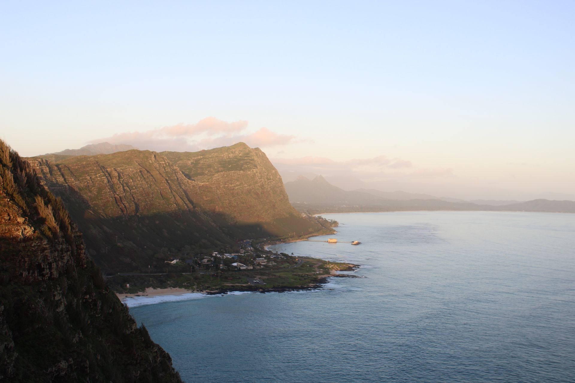 Rocky coastal cliffs overlooking a calm blue ocean at sunset, with hazy mountains in the distance
