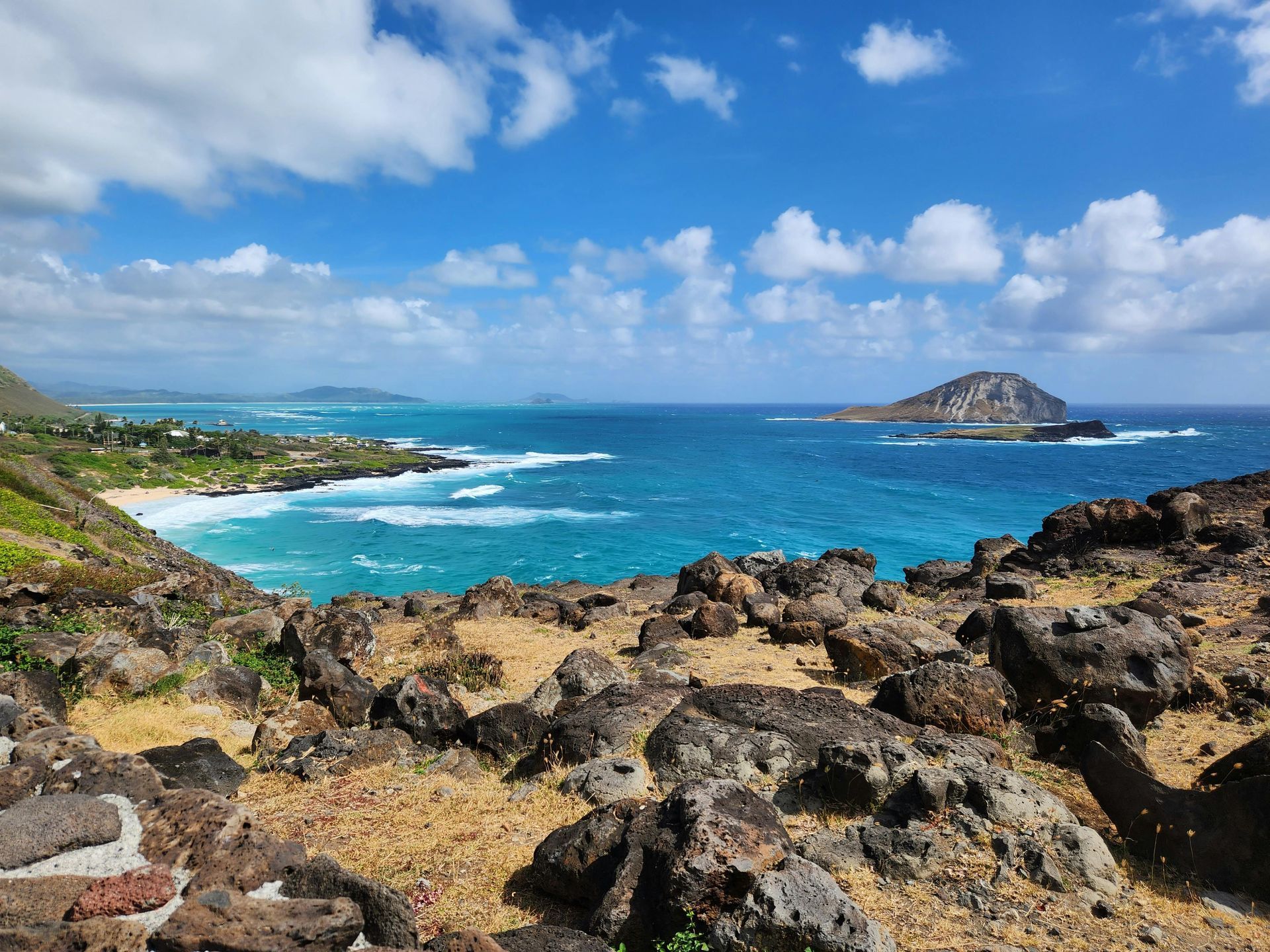 Rocky shoreline with turquoise ocean, waves, and a small island under a bright blue sky