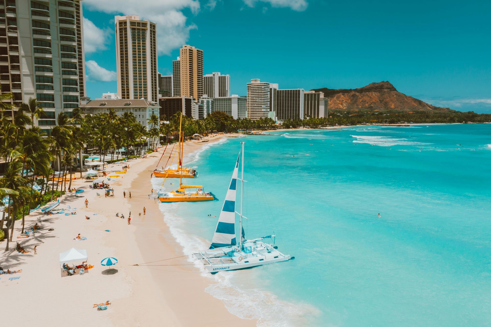 White-sand beach with turquoise water, sailboats, and tall beachfront hotels beside a green mountain.