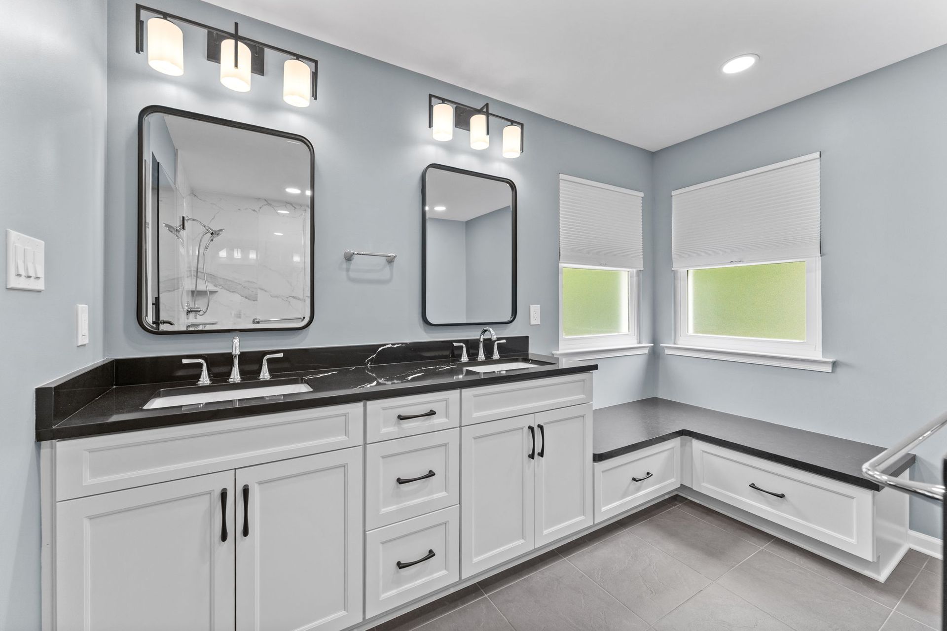 Bright modern bathroom with white vanity, black countertop, dual mirrors, and windows with green shades In Bucks County PA