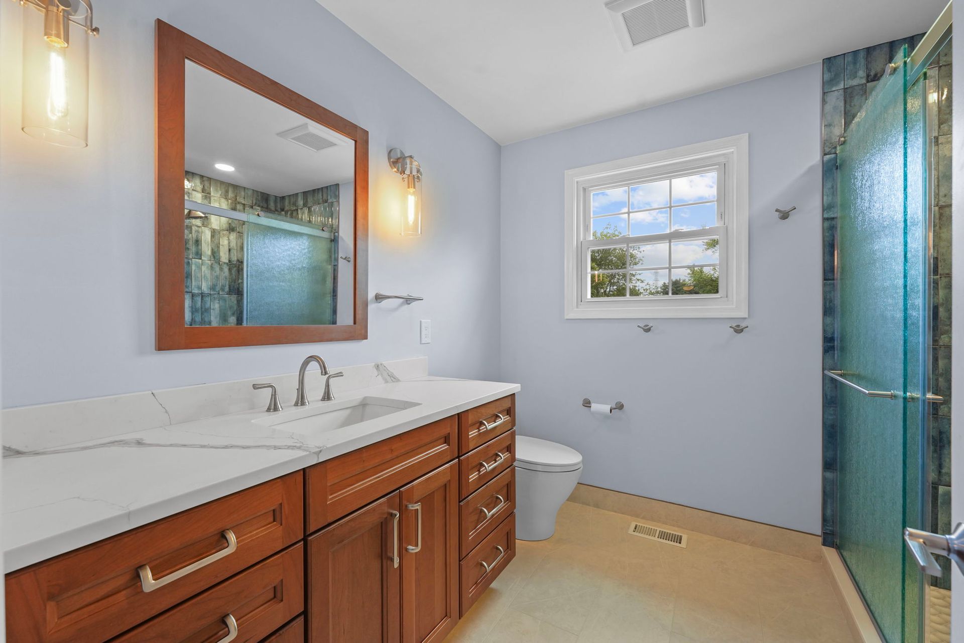 Bright bathroom with wooden vanity, white countertop, window, and glass-enclosed shower  In Bucks County PA