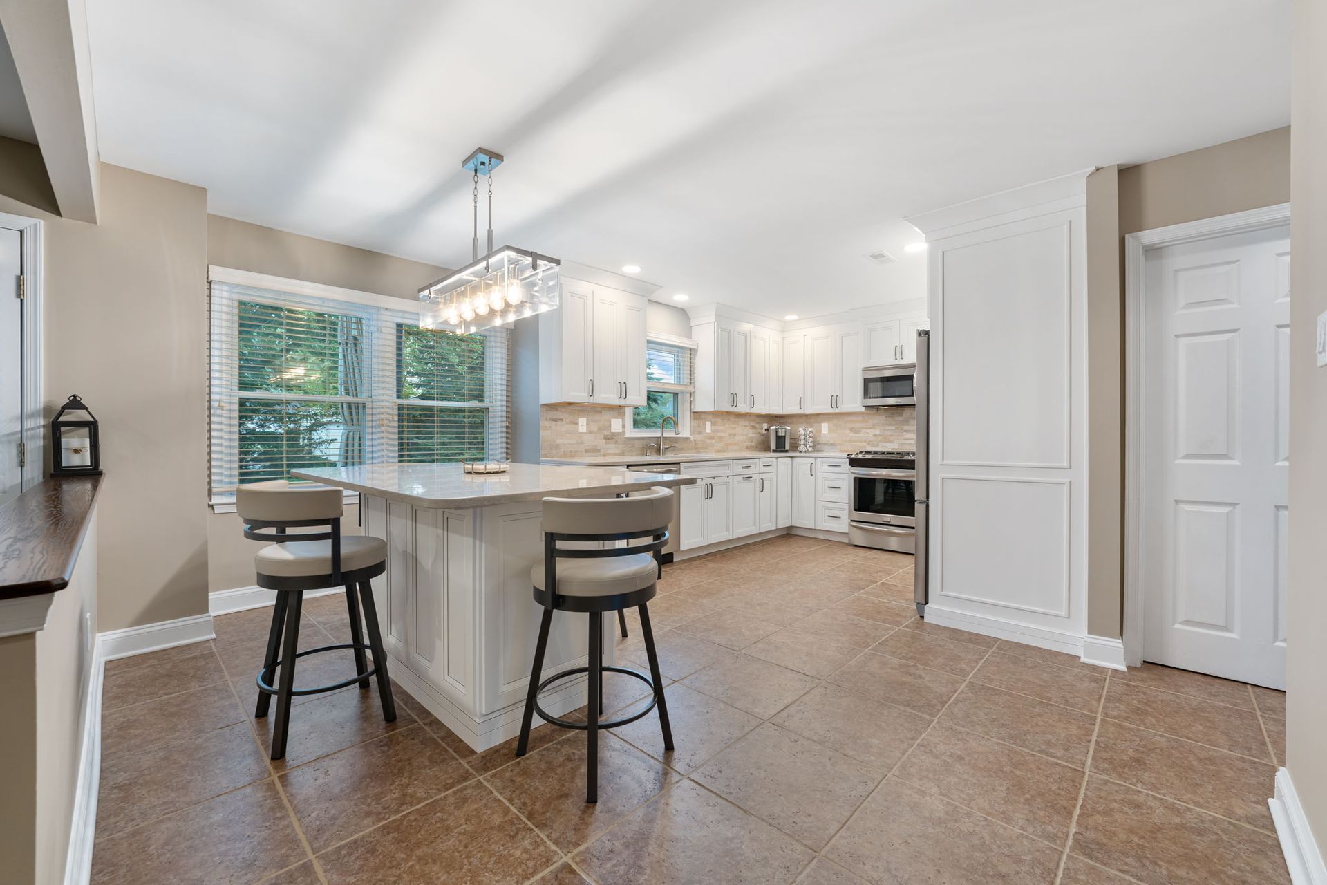 Bright kitchen with island, white cabinets, tan tile floor, and two stools near a large window In Bucks County PA