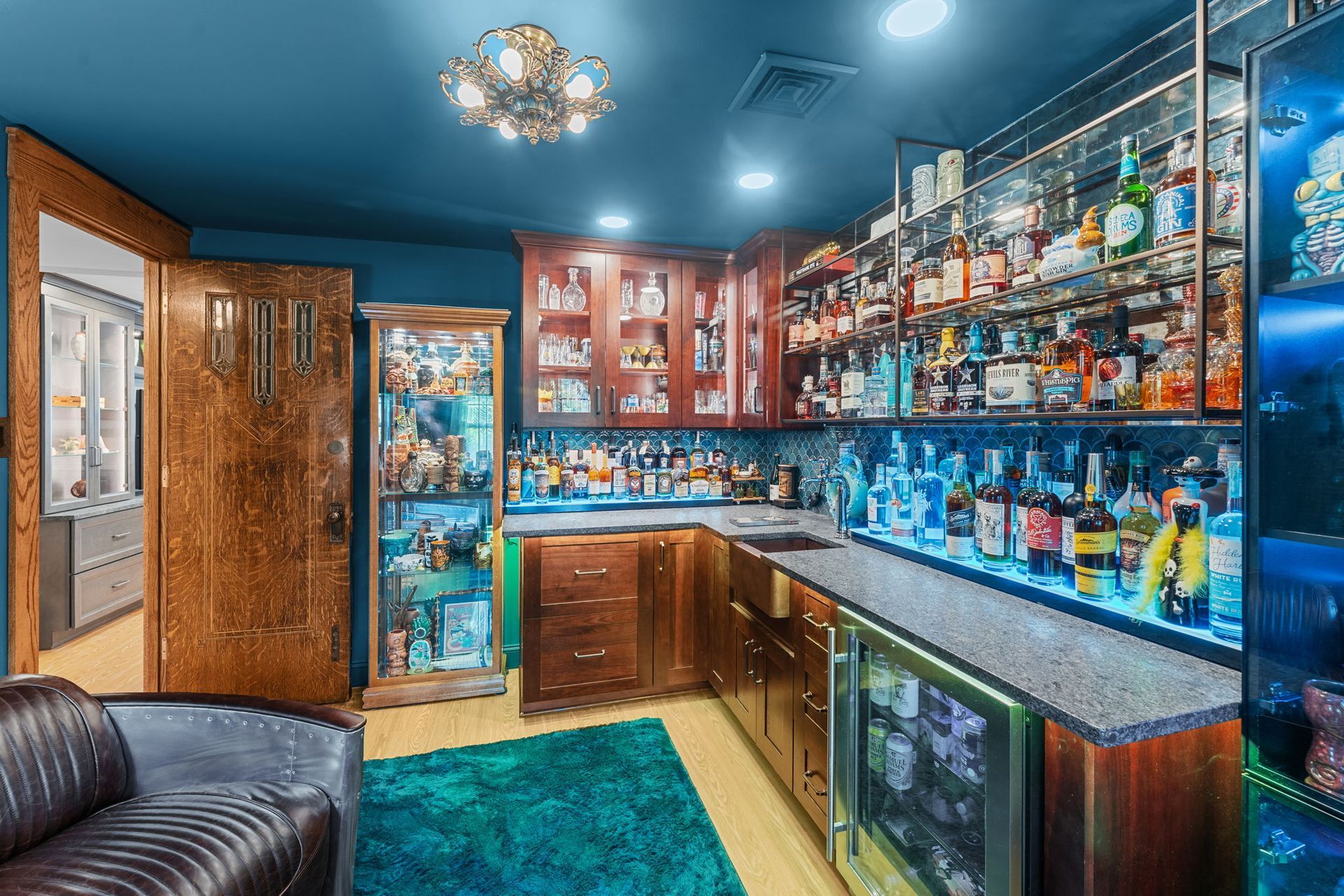 Colorful home bar with wood cabinets, shelves of bottles, and a black leather bench on a teal rug In Bucks County PA