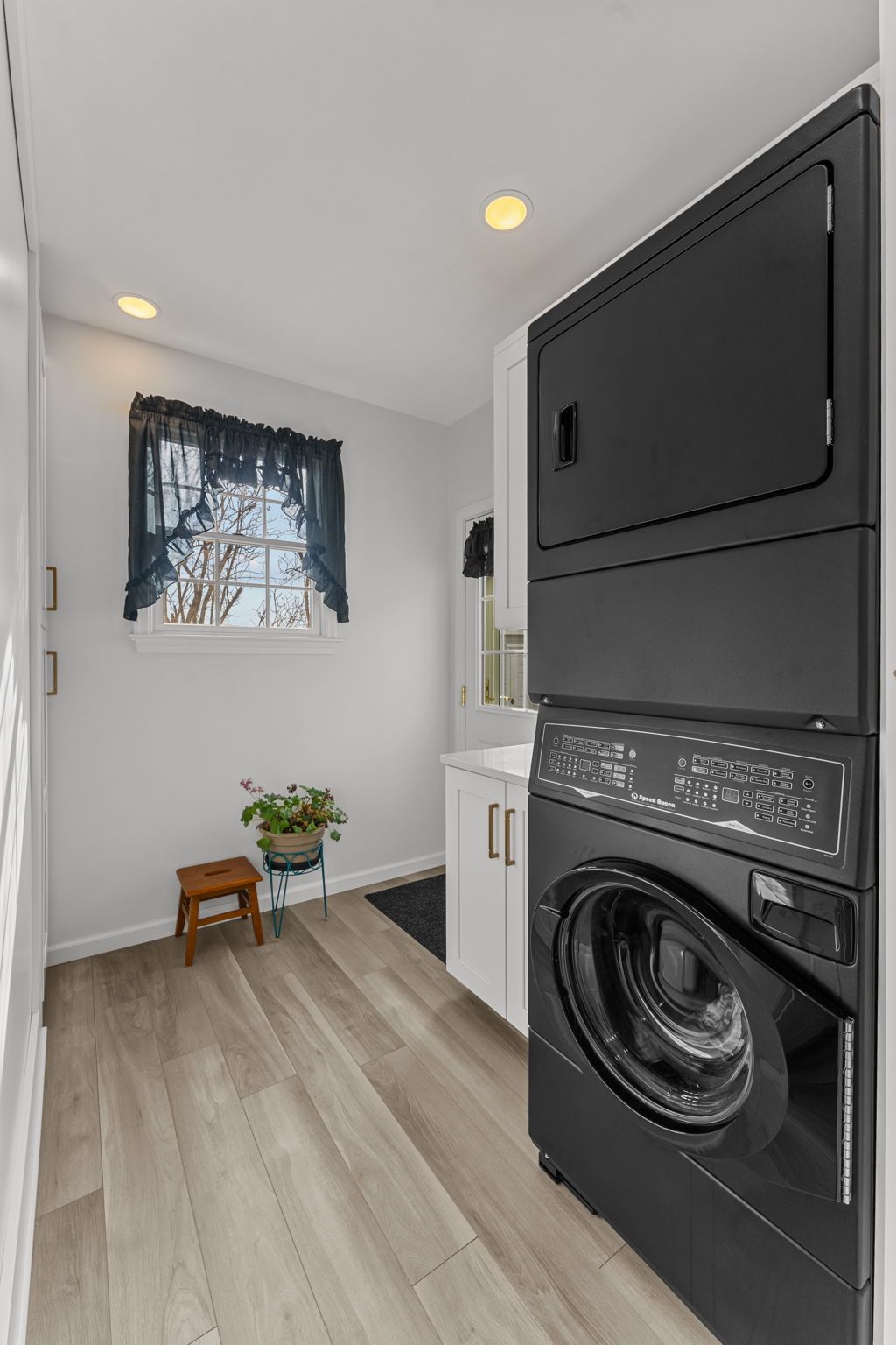 Compact laundry room with stacked black washer-dryer, white cabinets, window, and small plant stool In Bucks County PA