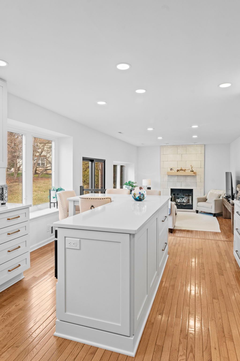Bright white modern kitchen with a large island, wood floors, and a fireplace in the adjacent living room In Bucks Country PA