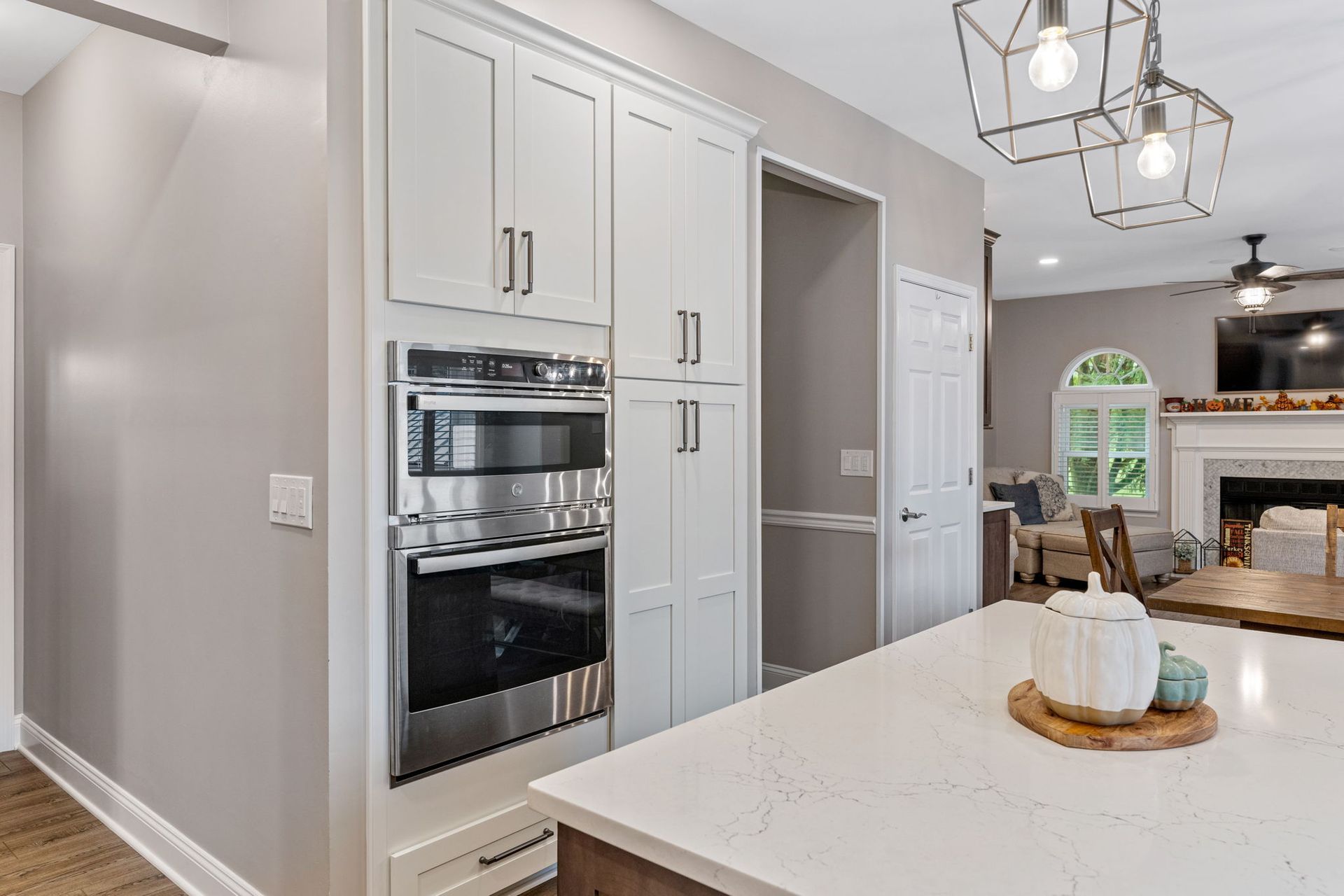Modern white kitchen with built-in oven, island, pendant lights, and fireplace in the background  In Bucks County PA