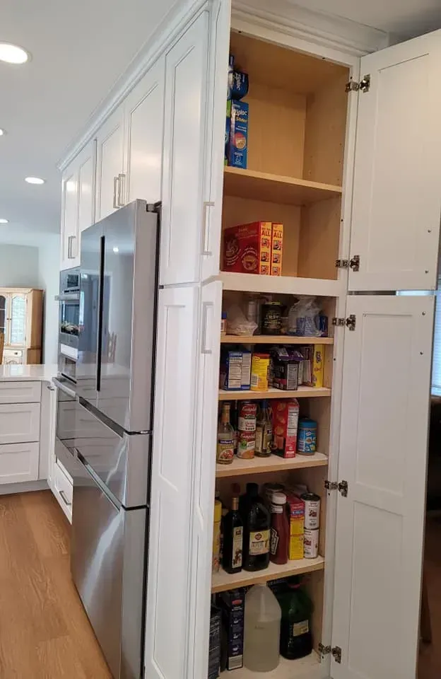 Open pantry cabinet beside a stainless steel refrigerator, stocked with food items in a white kitchen  In Bucks County PA