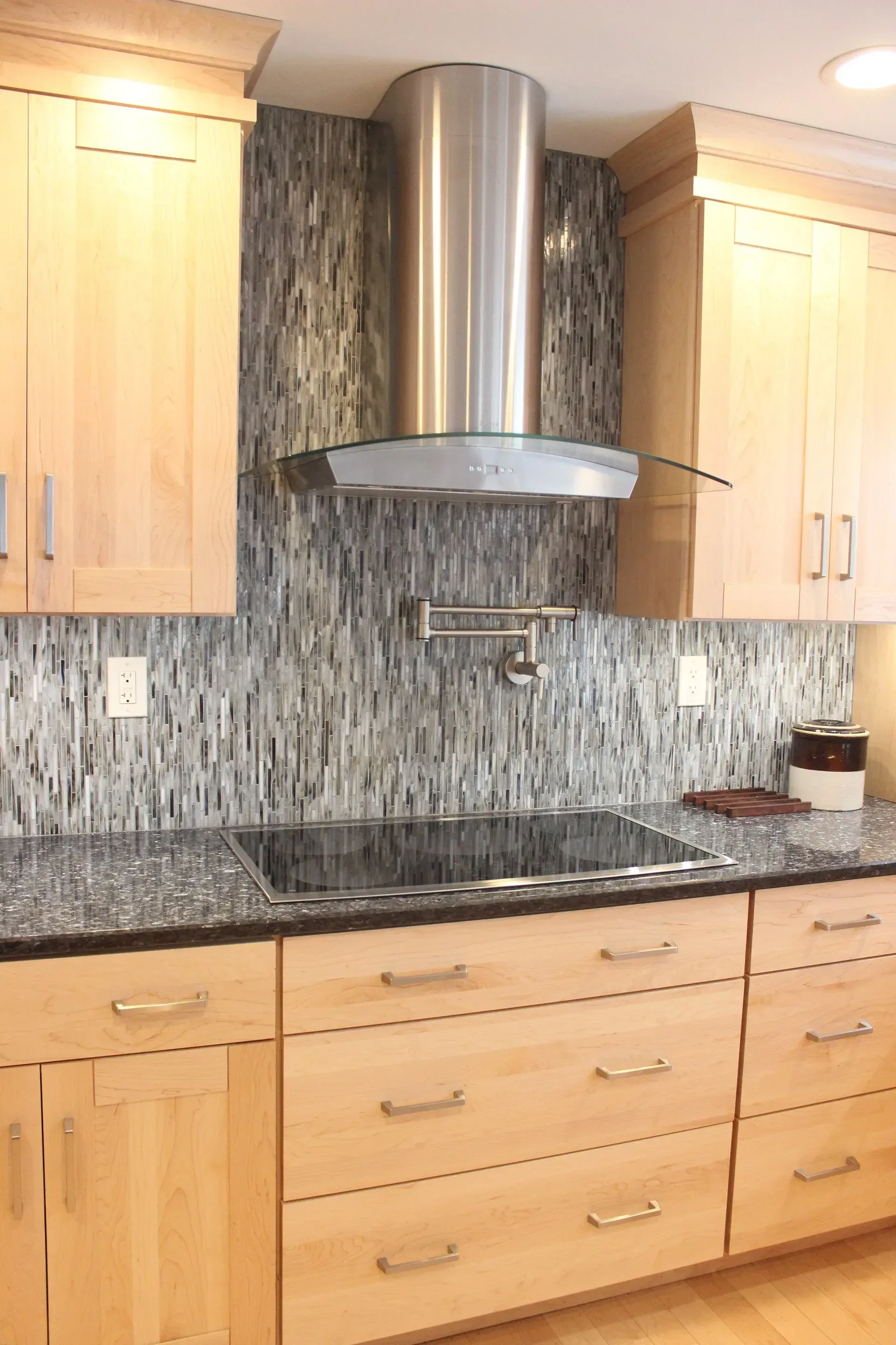 Modern kitchen with light wood cabinets, black countertops, and stainless steel range hood over a mosaic tile backsplash In Bucks Country PA