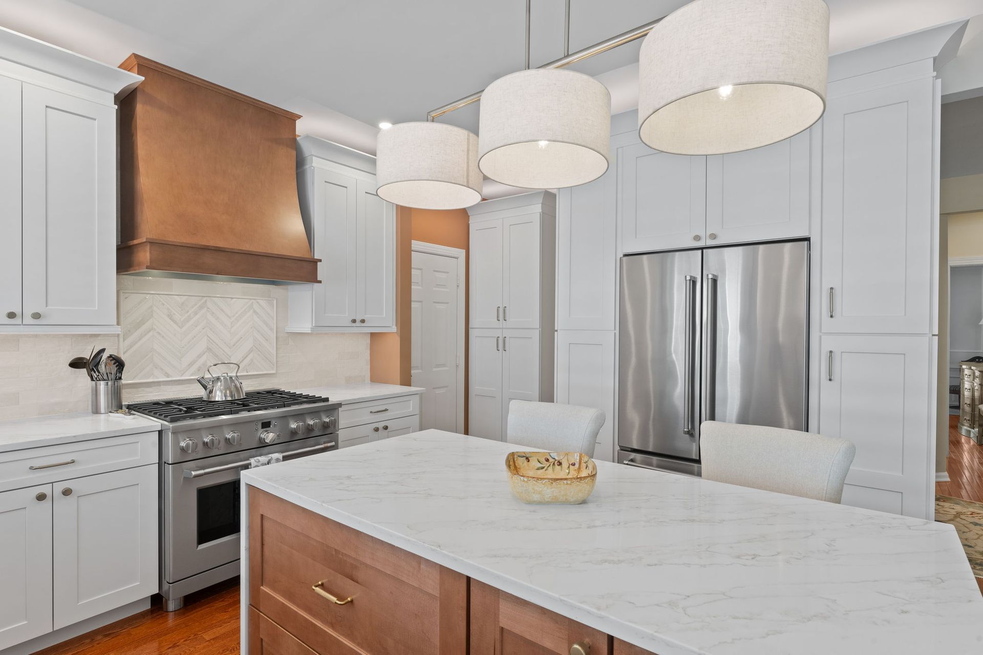 Modern white kitchen with stainless steel appliances, copper range hood, and marble island under pendant lights In Bucks County PA