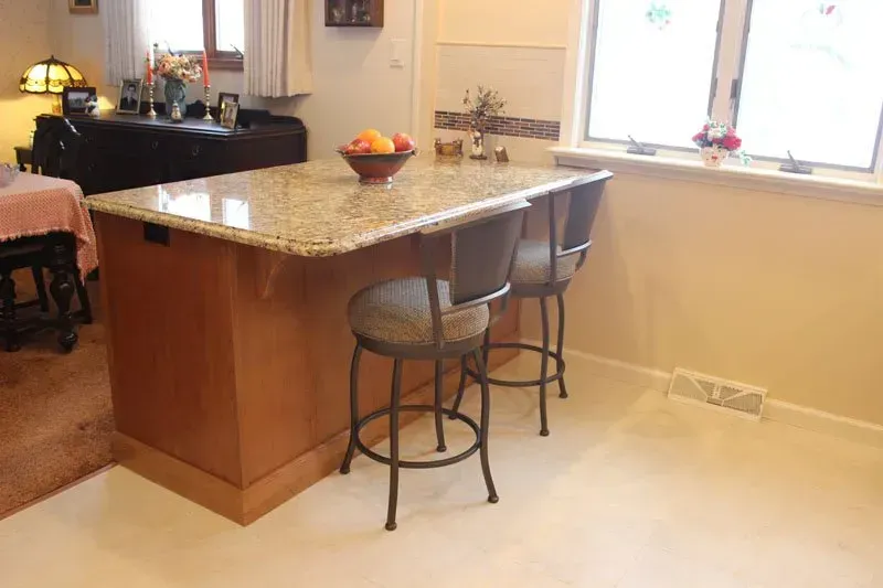 Kitchen with granite island, two bar stools, fruit bowl, and bright window over a light tile floor  In Bucks County PA
