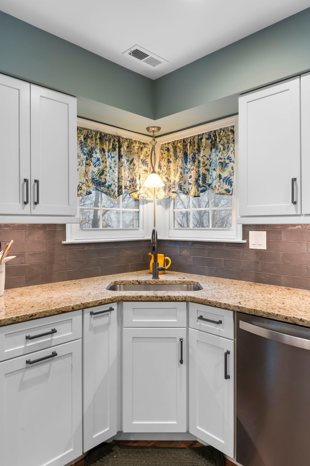 Small kitchen corner with white cabinets, speckled countertops, and a window over the sink with floral curtains In Bucks Country PA