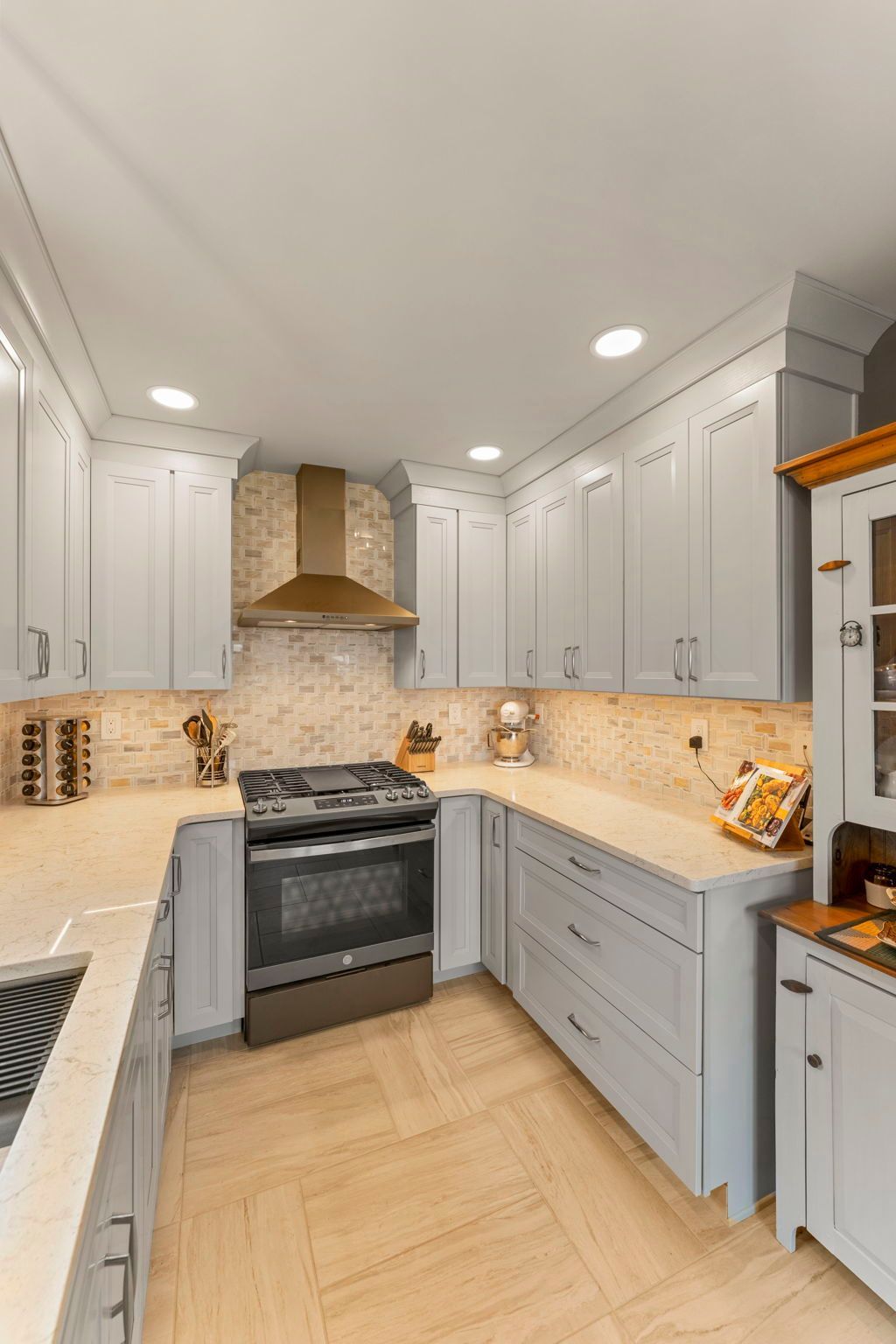 Bright narrow kitchen with white cabinets, beige tile floor, wood range hood, and stainless stove In Bucks County PA