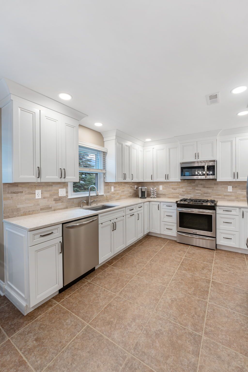 Bright modern kitchen with white cabinets, stainless appliances, beige tile floor, and large window above the sink In Bucks County PA