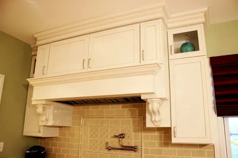 White kitchen cabinets above tiled backsplash and stove hood, with a small shelf and window blinds In Bucks County PA