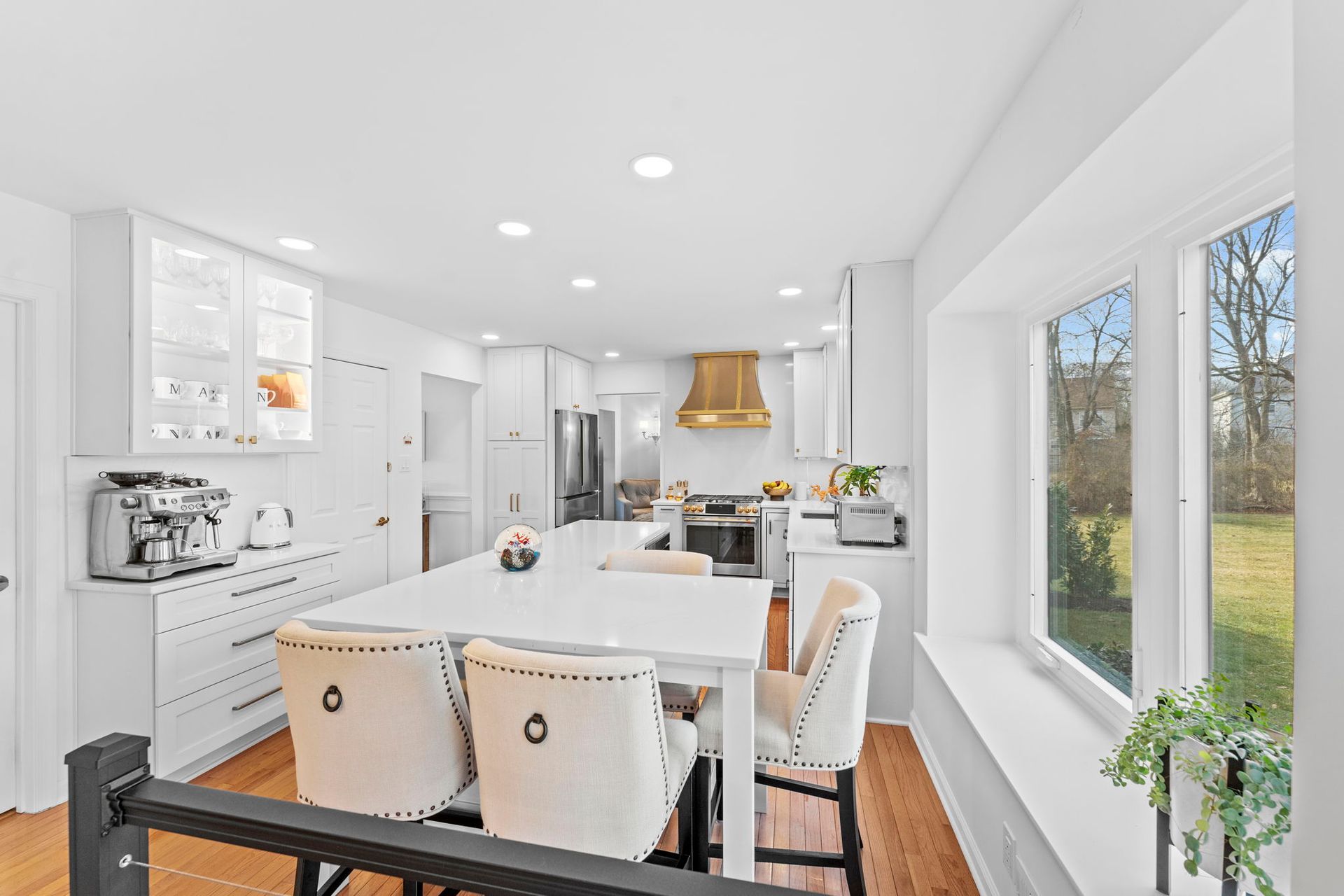 Bright white kitchen and dining area with island, stools, gold hood, and large windows overlooking a yard In Bucks Country PA