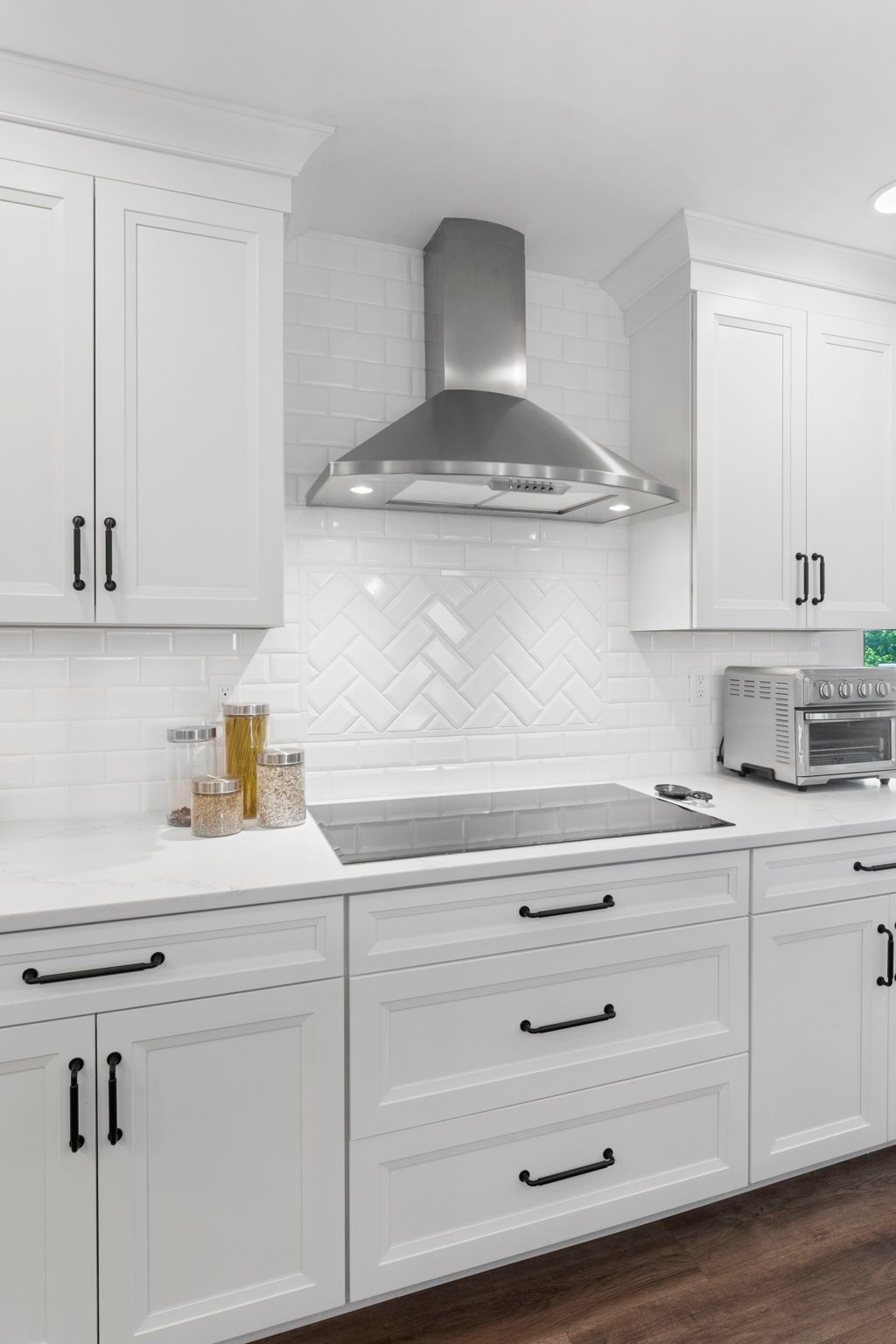 Bright white kitchen with stainless steel range hood, herringbone backsplash, and dark wood floor In Bucks County PA