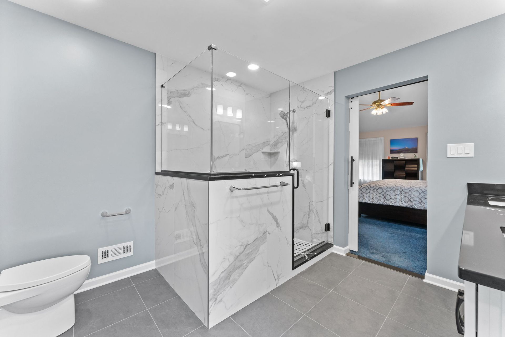 Modern gray bathroom with glass shower, white marble tile, and open doorway to bedroom In Bucks County PA