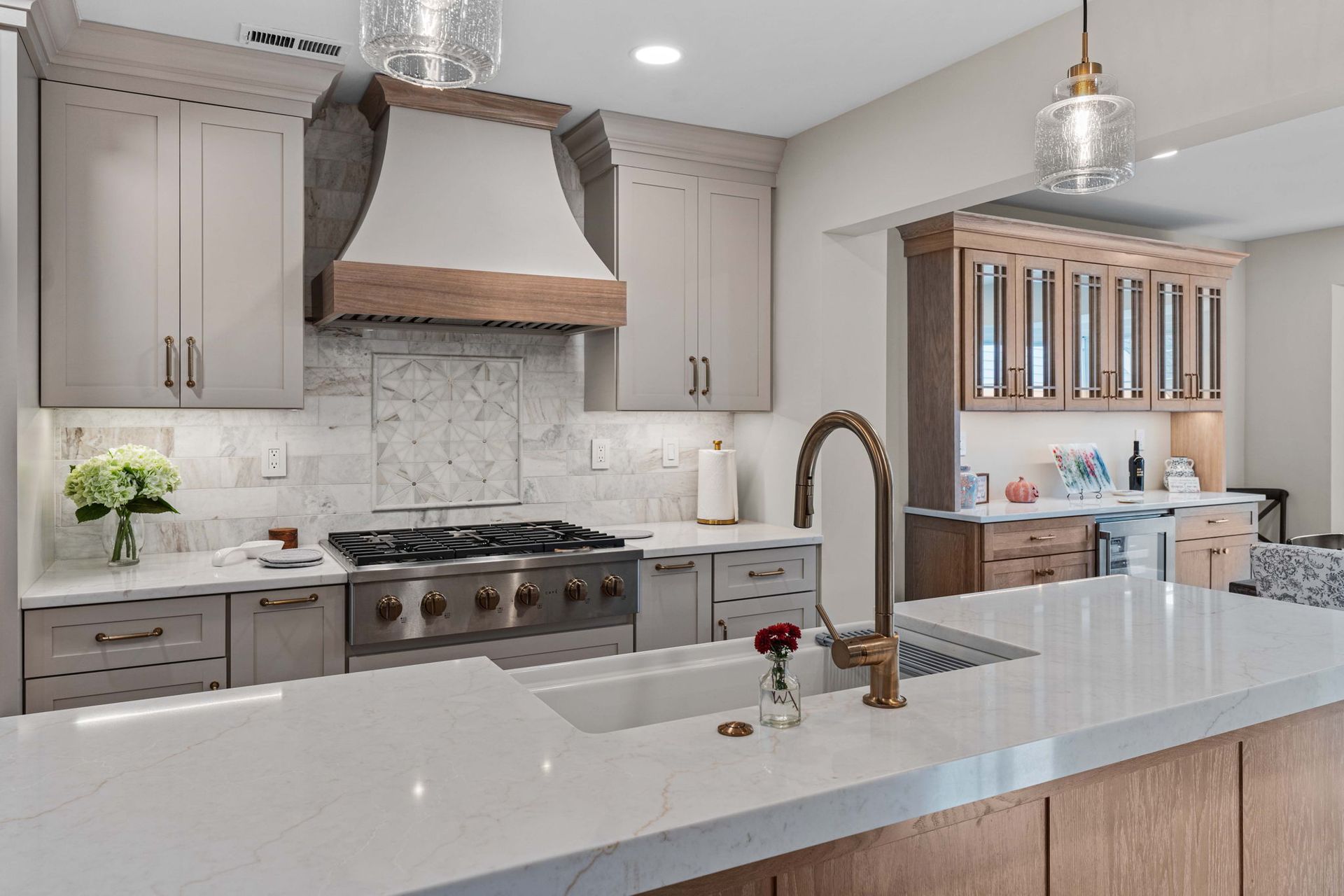 Elegant kitchen with taupe cabinets, a wood-accented range hood, marble backsplash, and a gold faucet over an island sink
