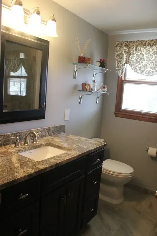 Small bathroom with dark vanity, granite sink, toilet, mirror, and window with patterned blind In Bucks County PA