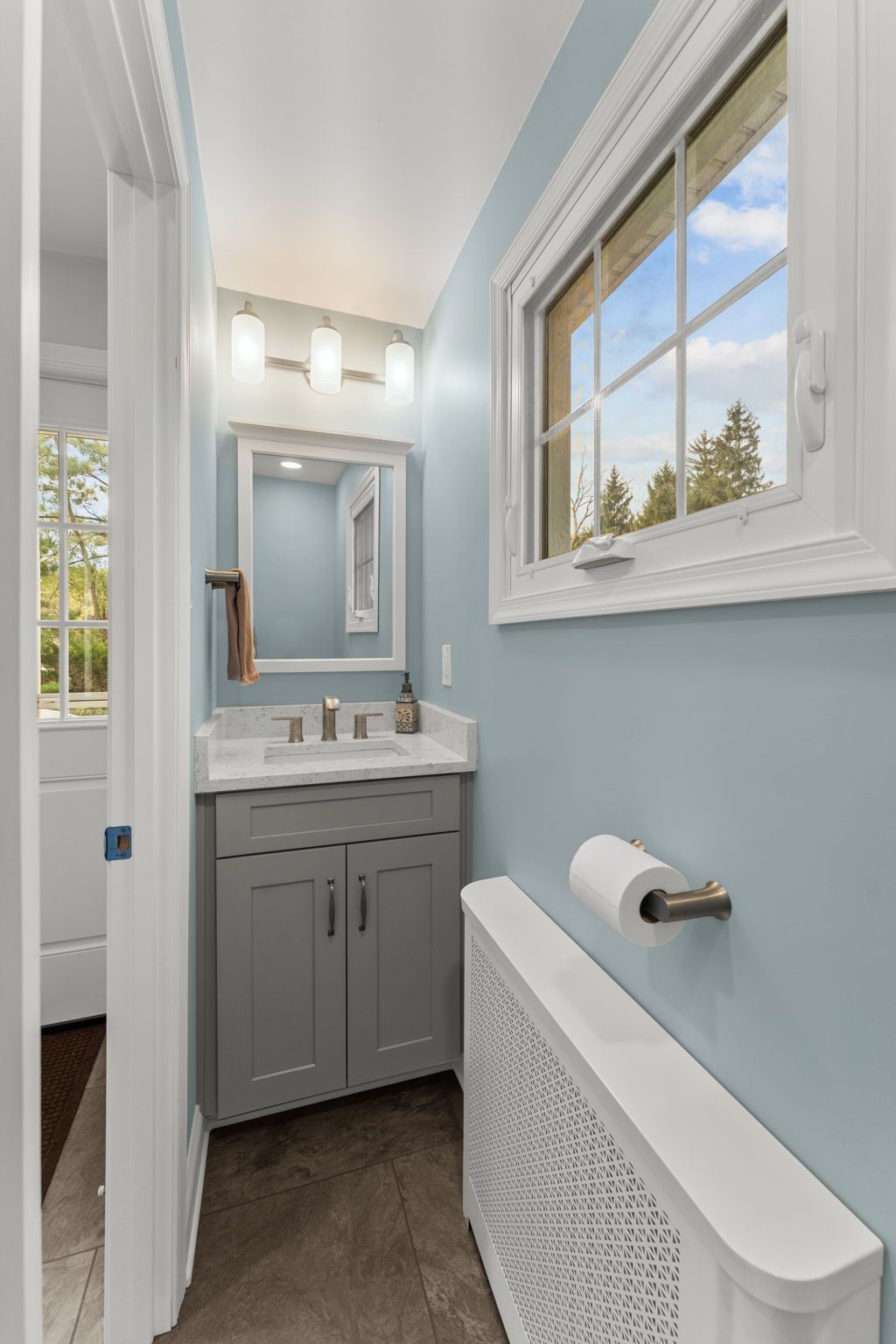 Narrow bathroom with gray vanity, light blue walls, white trim, and a window above a radiator  In Bucks County PA