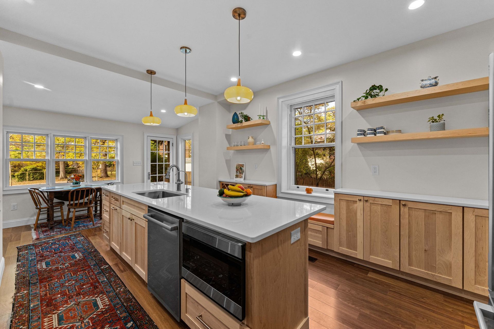 Modern kitchen with island, light wood cabinets, white counters, hanging pendant lights, and a dining area by windows In Bucks County PA