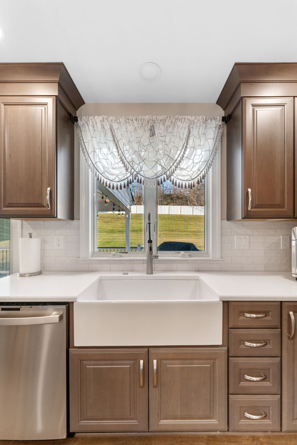 Kitchen sink under a window with lace curtains, flanked by brown cabinets and stainless dishwasher In Bucks Country PA