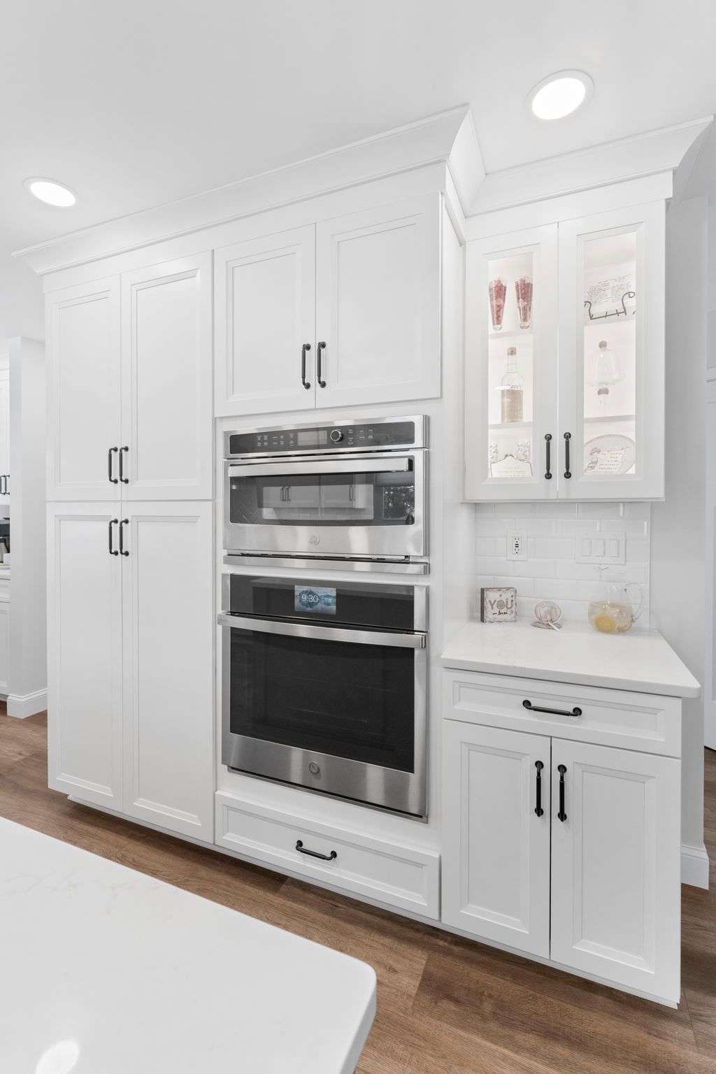 White built-in kitchen wall with stacked ovens, cabinets, and a small counter on wood flooring In Bucks County PA