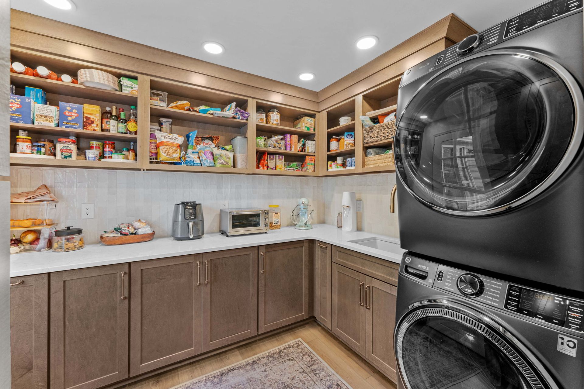 Laundry room with stacked black washer and dryer, white counters, wood cabinets, and open pantry shelves with supplies in Bucks County PA
